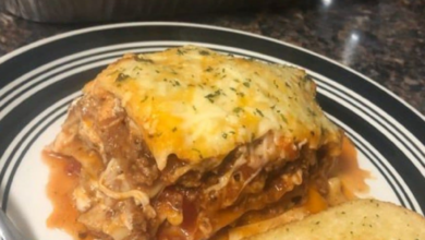 A generous serving of lasagna on a black and white plate, accompanied by a slice of garlic bread. The lasagna layers are visibly filled with meat, sauce, and a blend of melted cheeses, topped with a sprinkle of herbs. In the background, a baking dish filled with the remaining lasagna is visible, indicating a freshly baked, golden, and bubbly top.