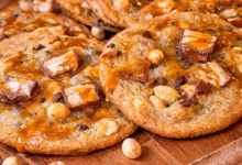 Bakery style Snickers cookies with gooey caramel, chocolate chunks, and peanuts on wooden board