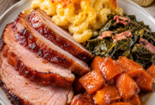 Honey glazed baked ham served with creamy mac and cheese, Southern collard greens, and candied sweet potatoes on a white dinner plate.