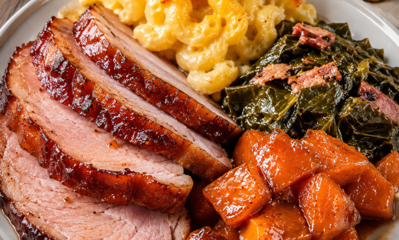 Honey glazed baked ham served with creamy mac and cheese, Southern collard greens, and candied sweet potatoes on a white dinner plate.